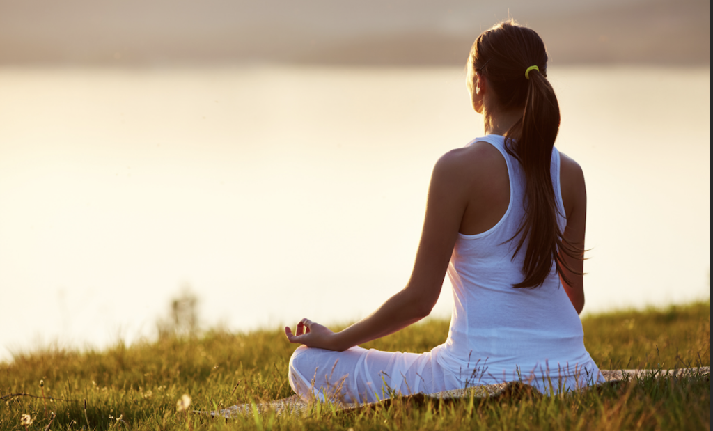 Woman meditating on a grassy hillside overlooking a body of water as part of her morning routine as a stay-at-home mom